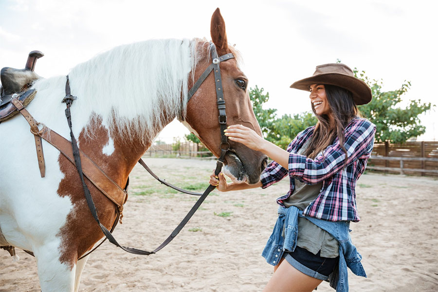 cheerful-charming-young-woman-cowgirl-with-horse-standing-laughing